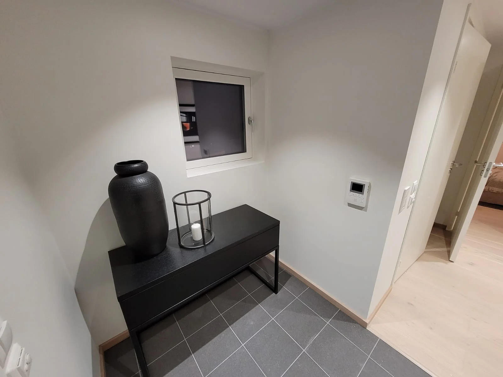 Entré med svart konsollbord, dekorvase og lykt på grå flislagt gulv etter renovering av leilighet